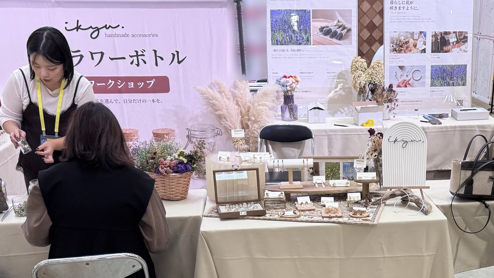 ikyuの「フラワーボトル ワークショップ」バナーの前で、スタッフが参加者に花材を手渡している。隣にはアクセサリーの展示テーブル