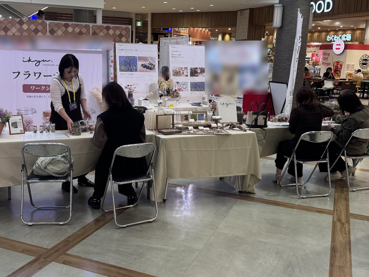 ikyuのバナー前の制作席で、スタッフが参加者に花材の瓶を手渡している。中央にはアクセサリーの展示テーブル