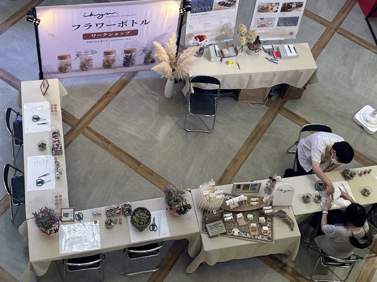 2階から見下ろしたikyuのブース。フラワーボトルのバナーと展示台、L字に並べた制作席で参加者が制作している