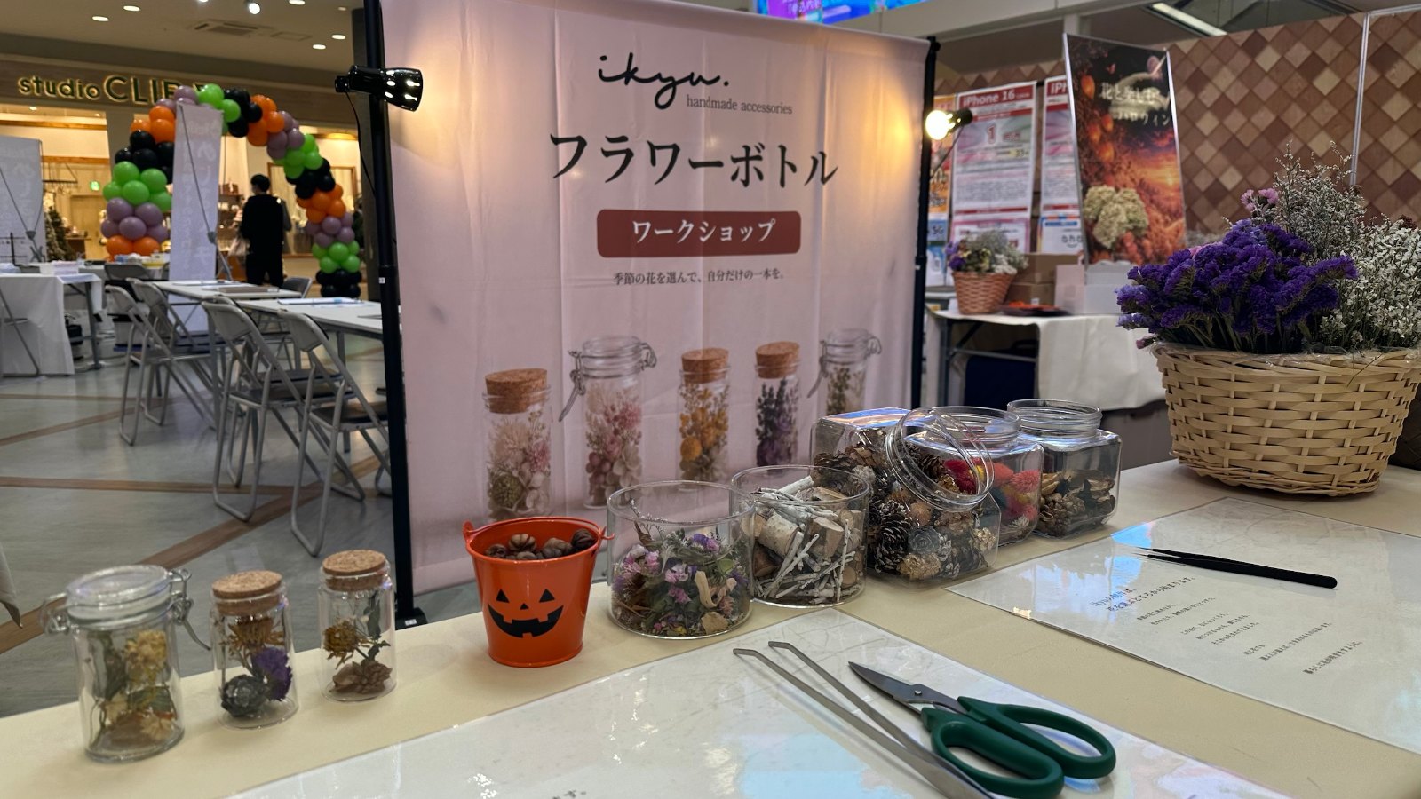 ikyuの「フラワーボトル ワークショップ」のバナーの前に、見本のフラワーボトルと花材の瓶、ハロウィンのかぼちゃのバケツ、ピンセットとハサミが並ぶ制作テーブル