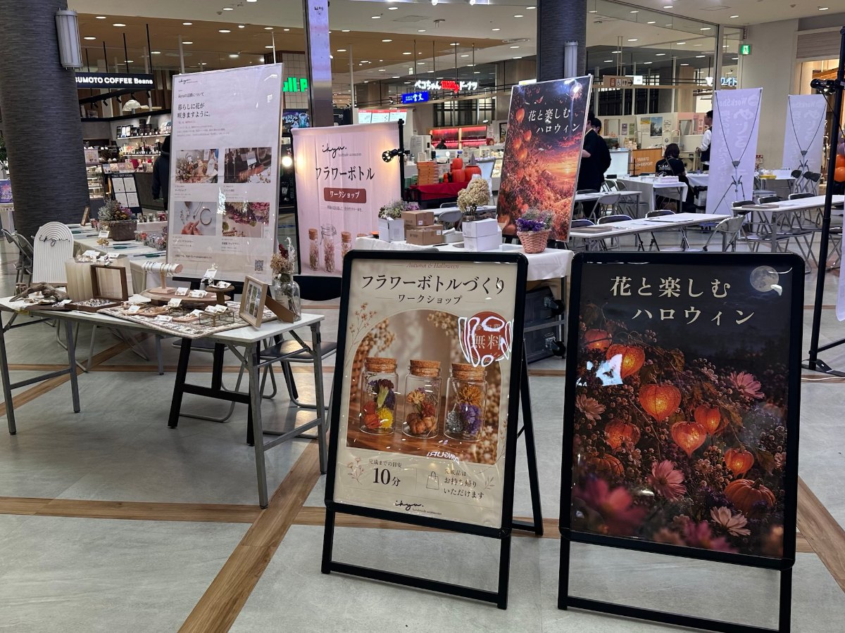 通路側に並ぶ「フラワーボトルづくり ワークショップ」と「花と楽しむハロウィン」の看板。奥にikyuのバナーとアクセサリーの展示テーブル