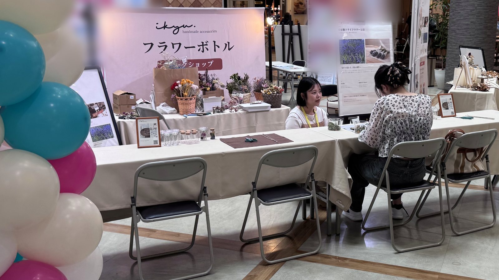 ikyuのフラワーボトルワークショップのブースで、作家がテーブル越しに参加者と向き合っている。左手前にはバルーン装飾、奥には企業イベントの会場が広がる