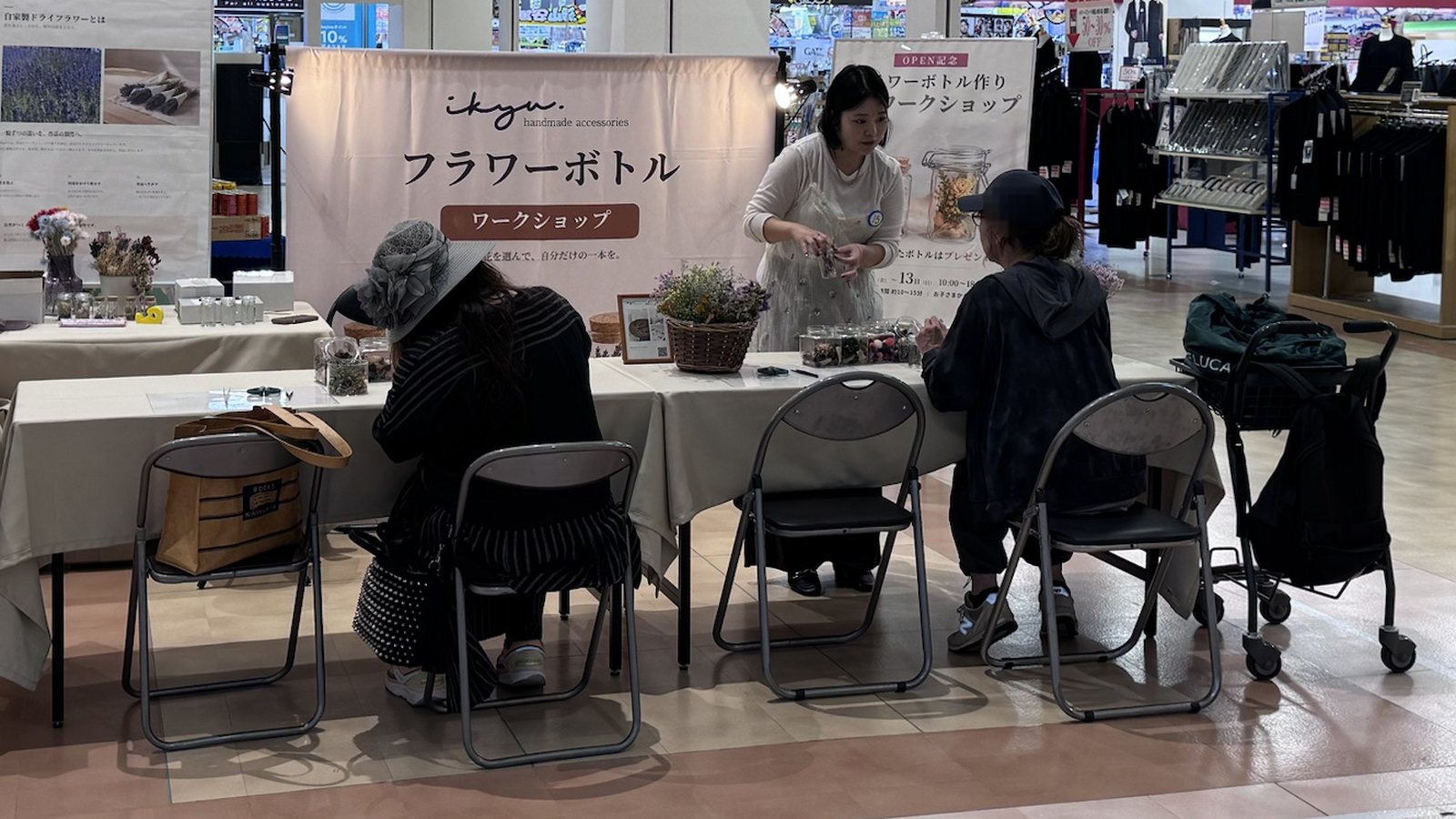 ikyuの「フラワーボトル ワークショップ」バナーの前で、参加者がテーブルに向かってフラワーボトルを制作し、スタッフが花材を手渡している
